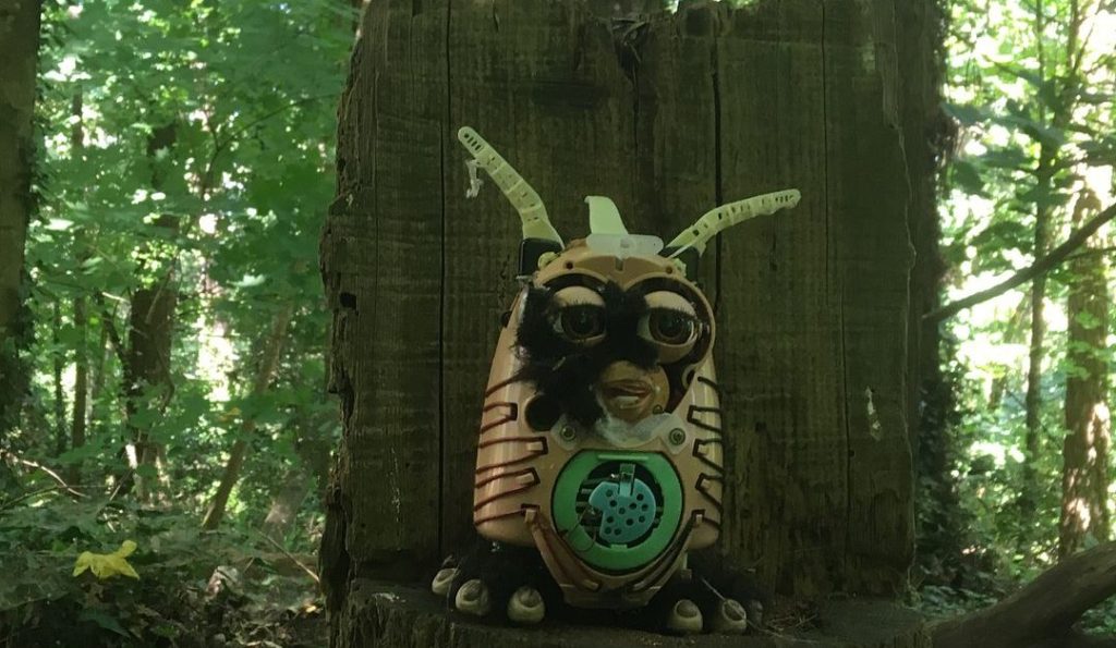 A mutilated Furby sits on an altar-like stump in the woods like some arcane woodland god